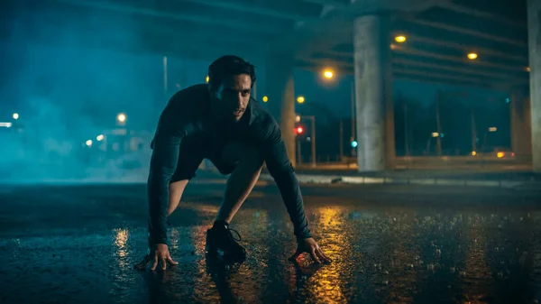 Strong Muscular Fit Young Man Προετοιμάζεται για Sprinting σε μια βροχερή βραδιά. Εκπαιδεύεται σε ένα αστικό περιβάλλον κάτω από μια γέφυρα με τα αυτοκίνητα στο παρασκήνιο. — Φωτογραφία Αρχείου