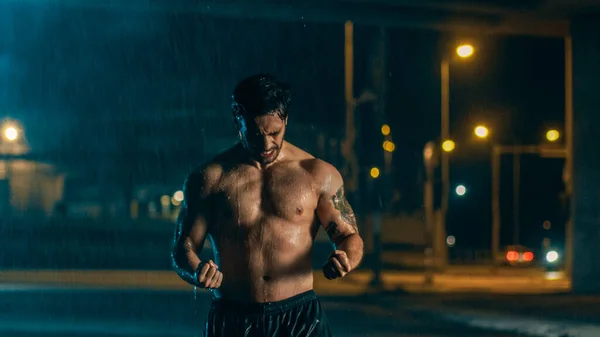Молодой спортсмен без рубашки празднует свои спортивные достижения в дождливую ночь и демонстрирует свои мускулы. Он в городской среде под мостом с автомобилями на заднем плане. — стоковое фото