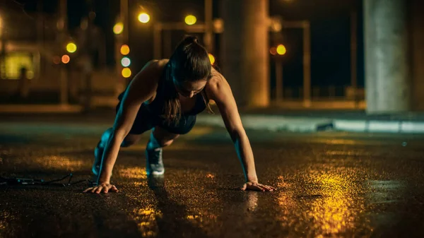 Bella Energetic Fitness Girl in Top atletico nero e pantaloncini sta facendo Push Up Esercizi. Sta facendo un allenamento in un ambiente urbano umido di sera sotto un ponte. — Foto Stock