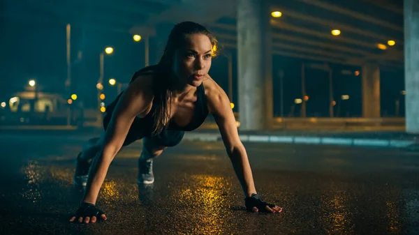 Gyönyörű Energetikai Fitness Girl fekete atlétikai Top és rövidnadrág csinál push up gyakorlatok. Edzést tart egy esténként nedves városi környezetben a híd alatt.. — Stock Fotó