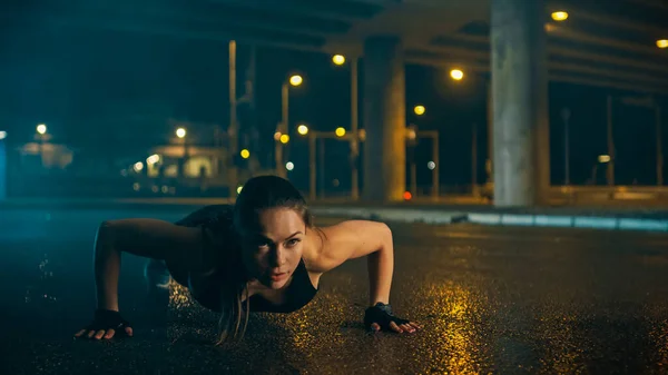 Beautiful Energetic Fitness Girl en camiseta y pantalones cortos deportivos negros está haciendo ejercicios push up. Ella está haciendo un entrenamiento en un ambiente urbano húmedo nocturno bajo un puente. —  Fotos de Stock