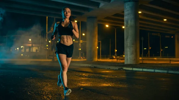 Hermosa chica de fitness en top atlético negro y pantalones cortos es trotar en la calle. Ella está haciendo un entrenamiento en un ambiente urbano húmedo nocturno bajo un puente. — Foto de Stock