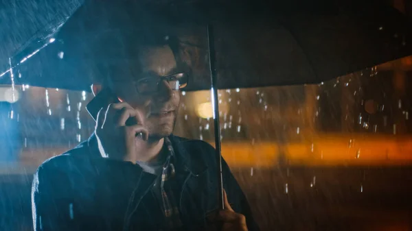 Νεαρός Καυκάσιος άνδρας με γυαλιά, φορώντας τζιν παλτό και τετράγωνο πουκάμισο μιλάει σε ένα τηλέφωνο κάτω από μια ομπρέλα. Είναι σκοτεινά έξω και βρέχει.. — Φωτογραφία Αρχείου