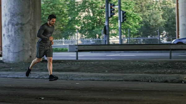 Athletic Young Man in Earphones and Sports Outfit is Jogging in the Street. Ele está correndo em um ambiente urbano sob uma ponte com carros no fundo. — Fotografia de Stock