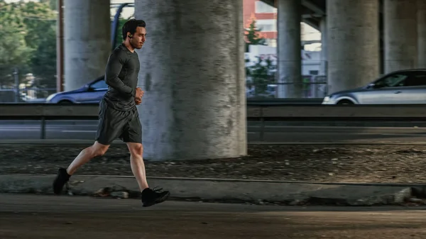 Athletic Young Man in Earphones and Sports Outfit is Jogging in the Street. Ele está correndo em um ambiente urbano sob uma ponte com carros no fundo. — Fotografia de Stock