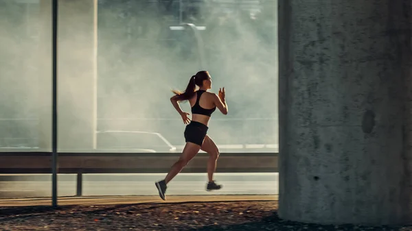 Девушка в черном спортивном топе и шортах энергично бежит по улице. Она бегает в городской среде под мостом с автомобилями на заднем плане. — стоковое фото