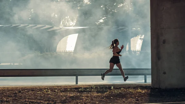 Gyönyörű Fitness Girl fekete atlétikai felső és rövidnadrág energetikusan fut az utcán. Egy városi környezetben kocog a híd alatt, autókkal a háttérben.. — Stock Fotó