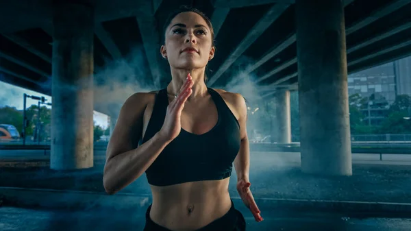 Retrato de una hermosa chica de fitness segura en top atlético negro y pantalones cortos corriendo a través de una calle ahumada. Se está ejecutando en un entorno urbano bajo un puente. — Foto de Stock