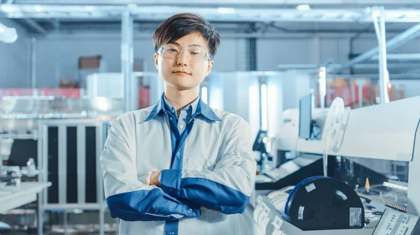 On High-Tech Factory: Portrait of Asian Worker with Crossed Arms. In the Background Electronic Printed Circuit Board Assembly Line that Uses Surface Mount Technology and Pick and Place Machinery.