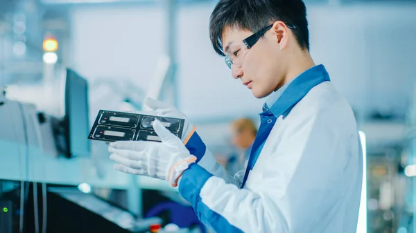 High-Tech Factory: Quality Control Engineer Checks Electronic Printed Circuit Board it for Damages. In the Background Assembly Line for PCB with Surface Mount Pick and Place Technology.