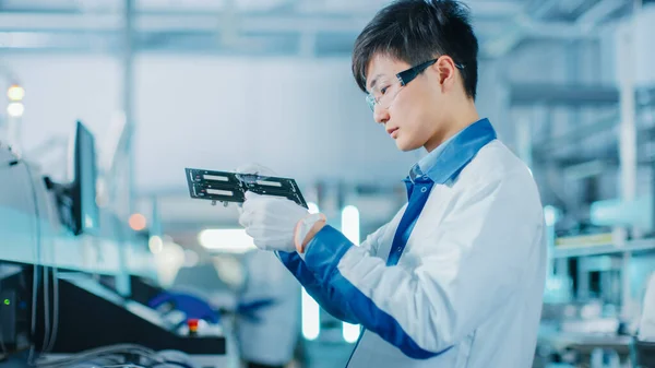 High-Tech Factory: Quality Control Engineer Checks Electronic Printed Circuit Board it for Damages. In the Background Assembly Line for PCB with Surface Mount Pick and Place Technology. — Stock Photo, Image
