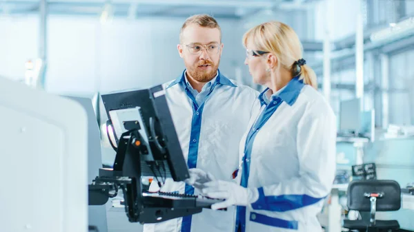 En la fábrica de alta tecnología, ingenieros femeninos y masculinos hablan y usan computadoras para programar máquinas electrónicas Pick and Place para la línea de ensamblaje de montaje en superficie de placa de circuito impreso. Producción de PCB con —  Fotos de Stock