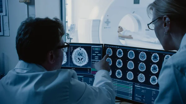 In Control Room Doctor and Radiologist Discuss Diagnosis while Watching Procedure and Monitors Showing Brain Scans Results, In the Background Patient Undergoes MRI or CT Scan Procedure. — Stock Photo, Image