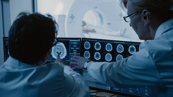 En la sala de control, el médico y el radiólogo discuten el diagnóstico mientras observan el procedimiento y los monitores que muestran los resultados de las exploraciones cerebrales, en el fondo, el paciente se somete a una resonancia magnética o a un procedimiento de exploración por TC. — Foto de Stock