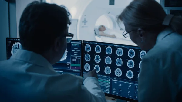 Врач и рентгенолог в диспетчерской обсуждают диагностику во время просмотра процедуры и мониторов, показывающих результаты сканирования мозга, в фоновом режиме пациент проходит МРТ или КТ. — стоковое фото