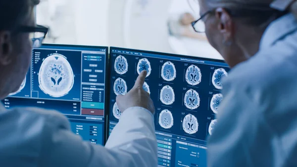 Врач и рентгенолог в диспетчерской обсуждают диагностику во время просмотра процедуры и мониторов, показывающих результаты сканирования мозга, в фоновом режиме пациент проходит МРТ или КТ. — стоковое фото
