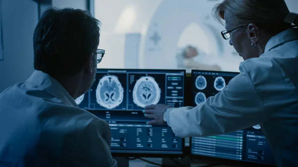 I kontrollrummet läkare och röntgenläkare Diskutera diagnos medan du tittar på procedur och monitorer visar hjärnskanning resultat, I bakgrunden patienten genomgår MRT eller datortomografi förfarande. — Stockfoto
