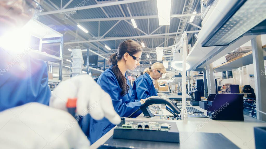 Female Electronics Factory Worker in Blue Work Coat and Protective Glasses is Assembling Laptops Motherboard with a Screwdriver. High Tech Factory Facility with Multiple Employees.