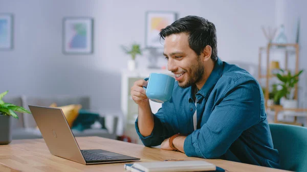 Πορτραίτο του χαμογελαστού νεαρού που εργάζεται σε ένα φορητό υπολογιστή από το σπίτι και πίνοντας καφέ από την κούπα και είναι ικανοποιημένη με το έργο του. Άνετο μοντέρνο επίπεδο γίνεται σε σκανδιναβικό στυλ. — Φωτογραφία Αρχείου