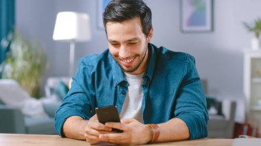 Yakışıklı Gülümseyen Adam oturma odasının masasında otururken akıllı telefon kullanıyor. Guy internette geziniyor, sosyal ağlar kullanıyor, evde dinleniyor..