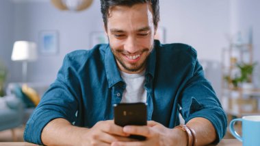 Yakışıklı Gülümseyen Adam Salonunda dinlenirken Akıllı Telefon kullanıyor. Guy internette geziniyor, sosyal ağları kullanıyor, evde eğleniyor..