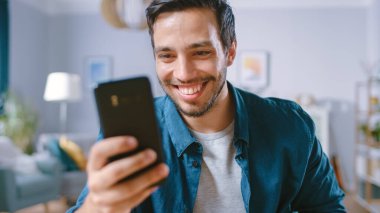 Yakışıklı Gülümseyen Adam Salonunda dinlenirken Akıllı Telefon kullanıyor. Guy internette geziniyor, sosyal ağları kullanıyor, evde eğleniyor..