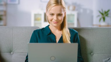 Güzel genç kadın evdeki bir koltukta rahatlıyor, dizüstü bilgisayar kullanıyor, interneti karıştırıyor, komik videolar izliyor ve e-alışveriş yapıyor..