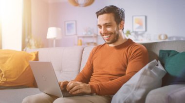 Evde oturan neşeli genç adam kucağında dizüstü bilgisayar tutuyor, interneti karıştırıyor, sosyal ağlar, e-alışveriş yapıyor. Dairesinde Sıcak Güneş Işığı.