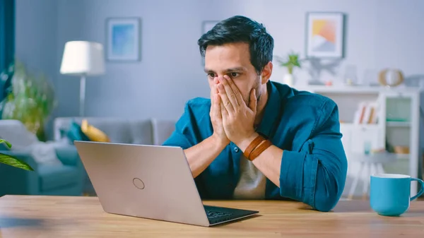 Skutt av en oppskaket ung mann dekker ansiktet med håndflater i Frustrasjon, mens han får dårlige nyheter fra å lese dårlige nyheter på en laptop. Opprørt mann hjemme. – stockfoto