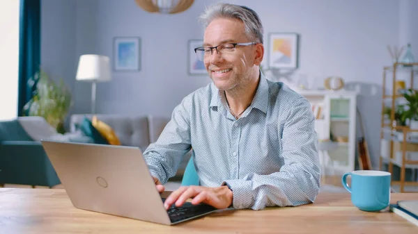 Όμορφος μεσήλικας χρησιμοποιεί φορητό υπολογιστή, ενώ κάθεται στο γραφείο του στο άνετο και κομψό σαλόνι. Ευτυχισμένος επιτυχημένος άνθρωπος εργάζεται στον υπολογιστή από το σπίτι. — Φωτογραφία Αρχείου