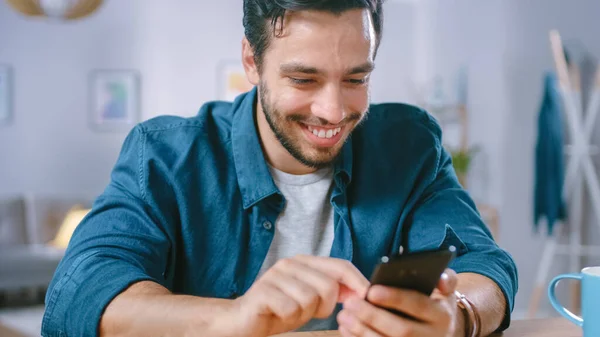 Schöner lächelnder Mann benutzt Smartphone, während er sich in seinem Wohnzimmer entspannt. Guy surft im Internet, nutzt soziale Netzwerke, hat Spaß zu Hause. — Stockfoto