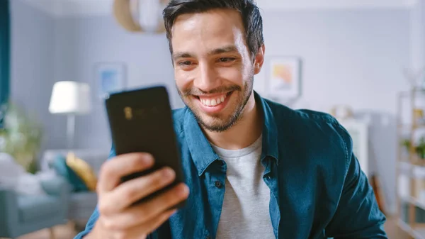 Bello uomo sorridente utilizza Smartphone mentre si rilassa nel suo salotto. Guy Navigare in Internet, Utilizzando i social network, Divertirsi a casa. — Foto Stock
