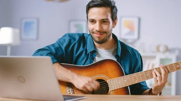 Utalentowany młody człowiek gra na gitarze siedząc przy biurku, czytając akordy z laptopa, robiąc blogi wideo online. Man Practicing gitara gra w domu. — Zdjęcie stockowe