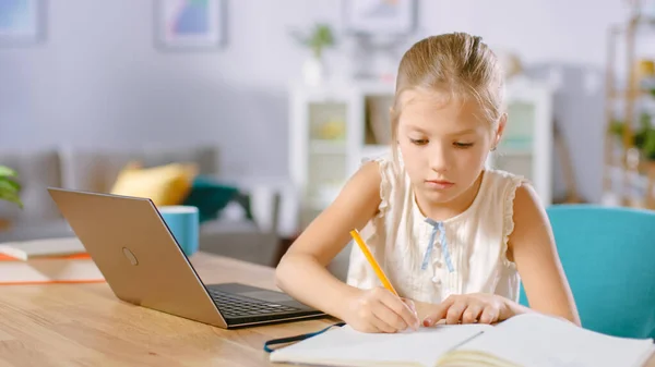 Smart Little Girl hace la tarea en su sala de estar. Ella sentada en su escritorio escribe con una pluma en sus libros de texto y utiliza el ordenador portátil. — Foto de Stock
