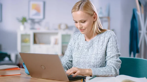 Vacker blond kvinna arbetar på en bärbar dator medan du sitter vid hennes skrivbord i vardagsrummet. Kvinnlig professionell frilansare som arbetar från mysiga hem. — Stockfoto