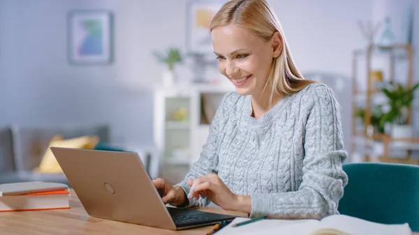 Vacker blond kvinna arbetar på en bärbar dator medan du sitter vid hennes skrivbord i vardagsrummet. Kvinnlig professionell frilansare som arbetar från mysiga hem. — Stockfoto