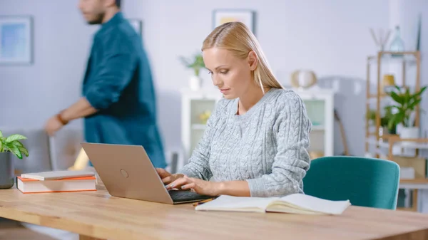 Gyönyörű nő dolgozik egy laptopon otthon, a barátja elsétál a háttérben. Professional Nő Freelancer Számítógép használata. — Stock Fotó