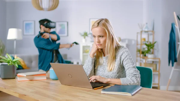 Mulher concentrada bonita trabalha em um laptop em sua mesa, atrás de seu namorado vestindo jogos de fone de ouvido de realidade virtual em jogo de vídeo com controladores — Fotografia de Stock