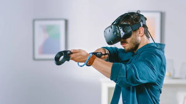 가상 현실 헤드셋을 착용하고 가정 비디오 게임에서 콘트롤러 게임을 하는 용감 한 청년. 거실에서 VR 슈터를 연주하는 사람. — 스톡 사진