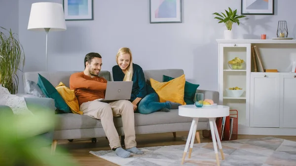 幸せな素敵なカップルは自宅でソファに座って、ボーイフレンドは膝の上にラップトップを保持し、インターネットを介して閲覧し、ストリーミングサービスを使用して電子ショッピングを行います。居心地の良いリビングルームで幸せな家族. — ストック写真