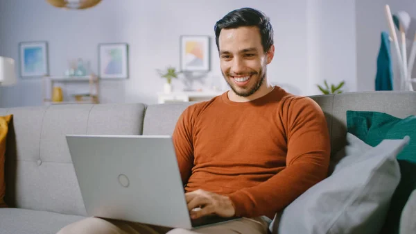 Cheerful Young Man at Home Sitting on a Sofa Holds Laptop on His Lap, Περιηγήσεις μέσω του Διαδικτύου, Κοινωνικά Δίκτυα, Κάνει e-shopping. — Φωτογραφία Αρχείου