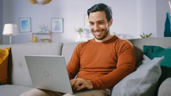 Cheerful Young Man at Home Sitting on a Sofa Holds Laptop on His Lap, Περιηγήσεις μέσω του Διαδικτύου, Κοινωνικά Δίκτυα, Κάνει e-shopping. — Φωτογραφία Αρχείου