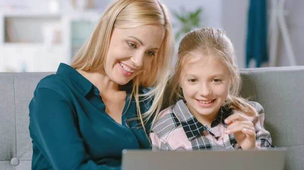 Hermosa joven mamá y su linda pequeña hija utilizar el ordenador portátil mientras se sienta en un sofá en casa. Familia pasar tiempo juntos viendo videos y dibujos animados en la computadora. — Foto de Stock