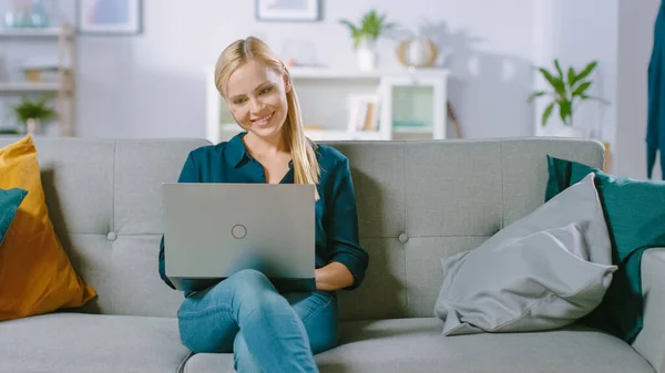 Hermosa mujer joven se relaja en un sofá en casa, Utiliza el ordenador portátil, Navega a través de Internet, Relojes Videos divertidos y hace e-Shopping. —  Fotos de Stock