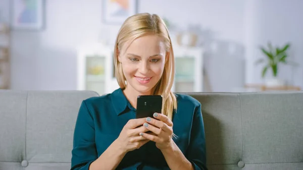Schöne blonde Frau benutzt Handy, während sie zu Hause auf einem Sofa sitzt. Glücklich lächelnde Frau nutzt Smartphone zum Surfen im Internet, in sozialen Netzwerken und zum Anschauen von Videos. — Stockfoto