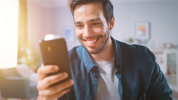 Schöner lächelnder Mann benutzt Smartphone, während er sich in seinem Wohnzimmer entspannt. Guy surft im Internet, nutzt soziale Netzwerke, hat Spaß zu Hause. Schuss in warmem Licht mit Sonneneruption. — Stockfoto