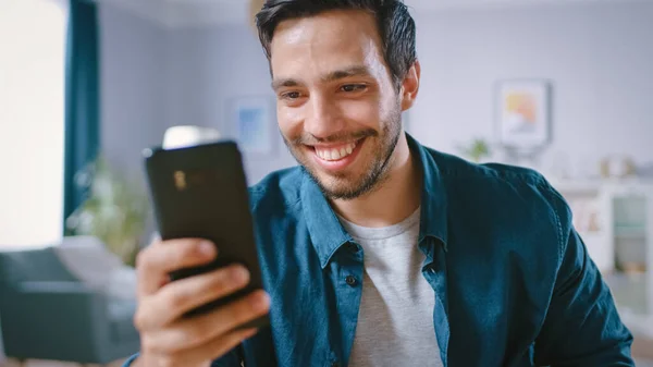 Schöner lächelnder Mann benutzt Smartphone, während er sich in seinem Wohnzimmer entspannt. Guy surft im Internet, nutzt soziale Netzwerke, hat Spaß zu Hause. — Stockfoto