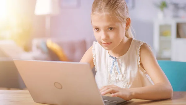 Симпатичная маленькая девочка пользуется ноутбуком, сидя за столом в гостиной. Ребенок занимается домашним хозяйством, просматривает Интернет и смотрит мультфильмы. Shot in Warm Light with Sun Flare. — стоковое фото