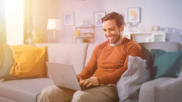 Jovem alegre sentado em um sofá segura laptop em seu lap, navega pela Internet, redes sociais, faz e-shopping. Homem em casa usando laptop enquanto sentado em um sofá. Luz quente do sol. — Fotografia de Stock