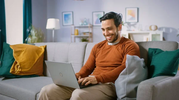 Vidám fiatalember ül egy kanapén tartja laptopján a Lap, Böngészések az interneten, a szociális hálózatok, nem e-Shopping. Férfi otthon Laptop használata, miközben ül a kanapén. — Stock Fotó